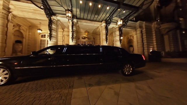 Video Reference: limousine, luxury car, black vehicle, driveway, hotel entrance, night, ornate architecture, covered entrance, columns, lanterns, transportation, exterior, celebration, arrival, formal