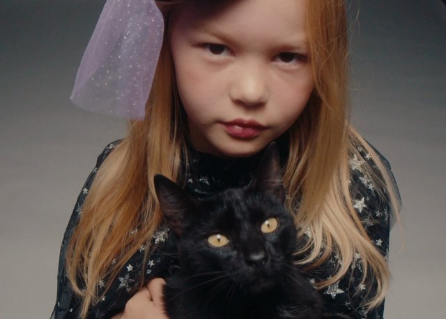 Video Reference: girl, child, black cat, portrait, close-up, pet, studio, gray background, blonde hair, star pattern, serious expression, animal, indoors, fashion, holding