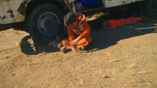 Video Reference: child, vehicle, wheel, roadside, sitting, lying down, outdoor, dusty, orange clothing, shadow, truck, accident scene, poverty, daytime