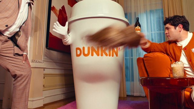 Video Reference: indoor, room, giant cup, costume, Dunkin, man, seated, iced drink, blur, motion, suit, orange cardigan, chair, curtains, wall art, advertisement