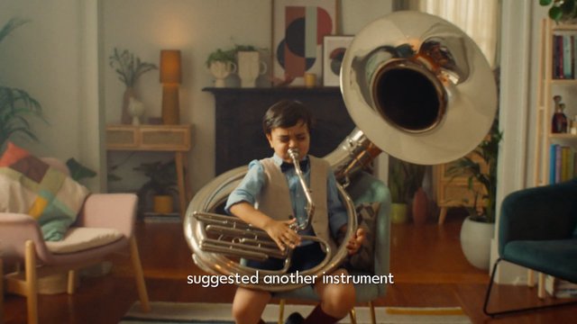 Video Reference: child, tuba, brass instrument, music, playing, indoor, living room, performance, home, instrument, young musician, couch, decor, warm lighting