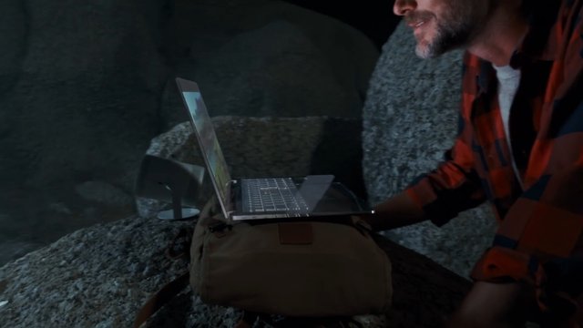 Video Reference: man, laptop, outdoor, rocky, night, technology, portable, working, plaid shirt, close-up, remote work, travel, adventure, low light