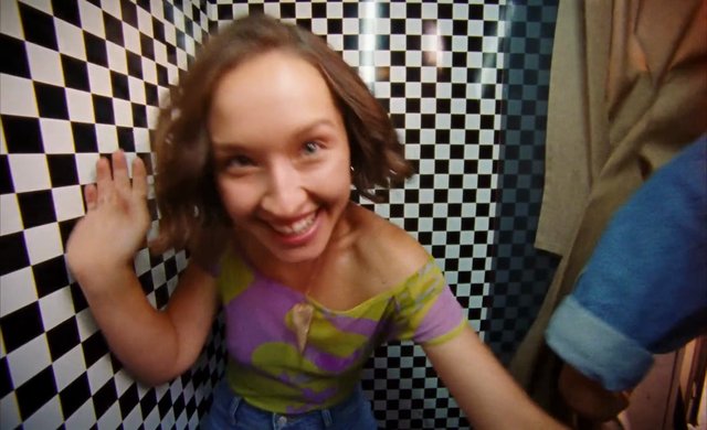 Video Reference: woman, smiling, portrait, indoor, checkered wall, wide angle, close up, colorful shirt, narrow space, selfie, casual, fashion