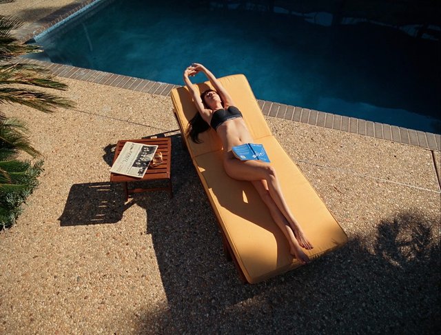 Video Reference: swimming pool, lounge chair, sunbathing, bikini, outdoor, relaxing, newspaper, drink, patio, summer, resort, poolside, stretching, woman