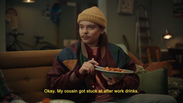 Video Reference: person, couch, living room, beanie, bowl of pasta, indoor, subtitles, casual clothing, snack, home interior, seated, conversation