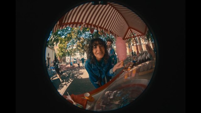Video Reference: fisheye, outdoor, cotton candy, street market, woman, canopy, plaza, park, fair, snack stand, sunlight, candid, vendor stall, people, summer, wide angle
