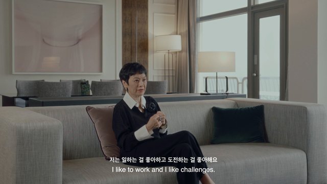 Video Reference: woman, interior, living room, sofa, interview, subtitles, modern home, sitting, conversation, indoor, elderly, furniture, home decor, window, lamp, portrait