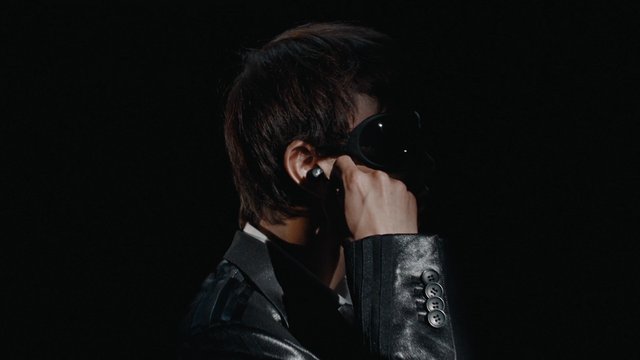 Video Reference: person, portrait, dark background, suit, sunglasses, visor, profile, fashion, studio lighting, close-up, hand gesture, black clothing
