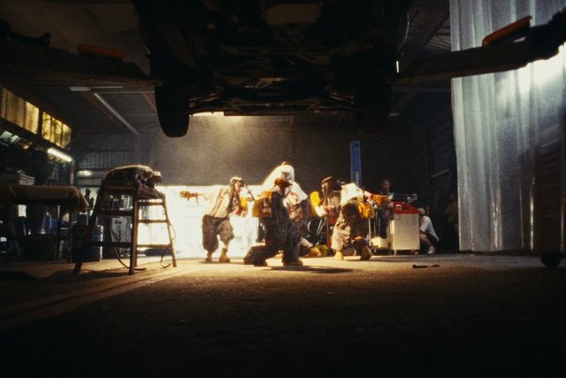 Video Reference: garage, workshop, industrial, people, crouching, tools, equipment, vehicle, underneath car, dimly lit, bright light, repair, maintenance, indoor, scene, clutter