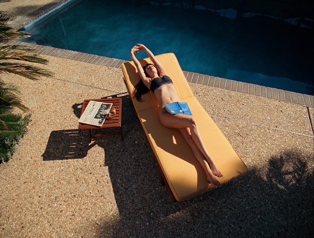 Video Reference: pool, lounge chair, sunbathing, woman, bikini, outdoor, summer, relaxation, swimming pool, sunlight, travel, resort, magazine, drink, leisure, backyard