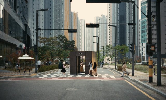 Video Reference: city street, crosswalk, pedestrians, high-rise buildings, urban neighborhood, traffic lights, street lamps, booth, sidewalk, trees, apartment towers, road, daytime, intersection, urban scene