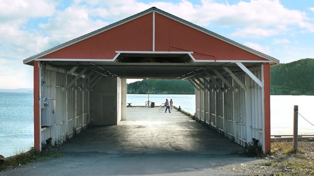 Video Reference: waterfront, boathouse, pier, dock, covered structure, red building, sea, bay, coastline, mountains, sky, outdoors, people, bicycle, landscape, harbor