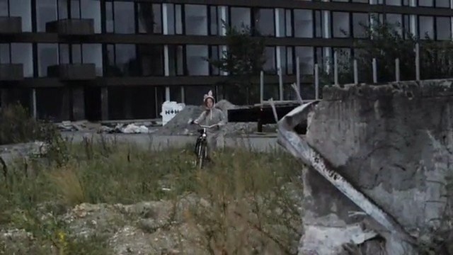Video Reference: person, bicycle, outdoors, abandoned building, ruins, concrete, grass, urban decay, rubble, glass facade, construction debris, overgrown, cycling, wasteland