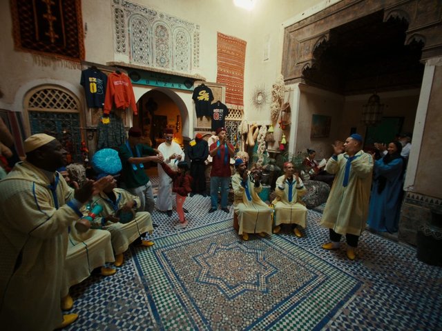 Video Reference: indoor courtyard, traditional clothing, music, dance, clapping, performance, ornate interior, tiled floor, arches, decorative walls, cultural celebration, group gathering, market stall, textiles, Moroccan style, tourist scene