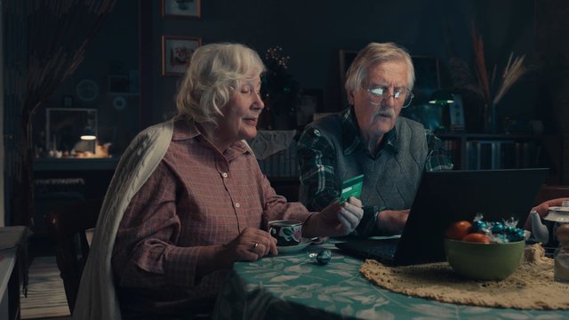 Video Reference: elderly couple, indoor, dining table, laptop, credit card, home, dim lighting, seniors, tableware, fruit bowl, candies, conversation, living room, casual clothing, household, technology