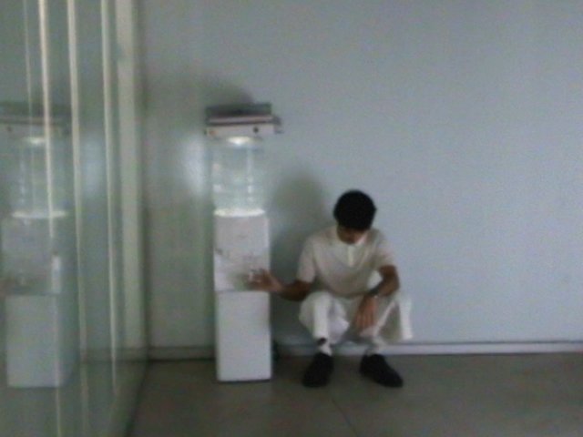 Video Reference: indoor, person, crouching, water dispenser, plain wall, minimal interior, blurry, standing water cooler, quiet room, white clothing, empty space