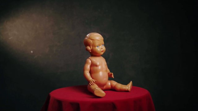 Video Reference: baby doll, toy, plastic, seated, red tablecloth, studio lighting, dark background, still life, object