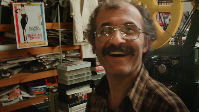Video Reference: man, smiling, glasses, mustache, storage room, workshop, shelves, tapes, poster, indoor, cluttered, close-up, casual, film, vintage