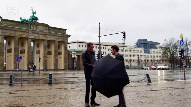 Video Reference: Sky, Building, Cloud, Window, Coat, Standing, Gesture, Road surface, Public space, Umbrella