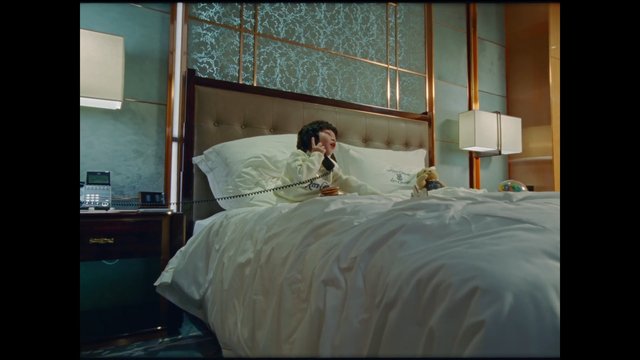 Video Reference: bedroom, hotel room, person, telephone, bed, indoor, nightstand, lamp, teddy bear, calling, interior, luxury, white bedding, sitting, cozy, modern