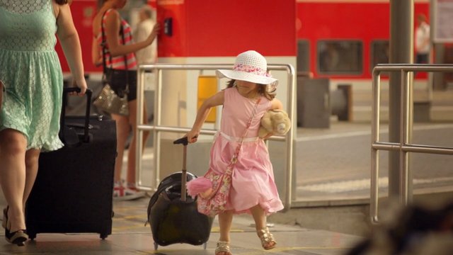 Video Reference: girl, child, train station, platform, suitcase, travel, pink dress, sunhat, teddy bear, commuting, family, train, outdoor, motion, travel luggage