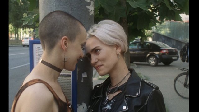 Video Reference: street, outdoor, two people, closeup, city, portrait, short hair, blonde hair, leather jacket, urban, tree, cars, bicycle, intimate, young adults