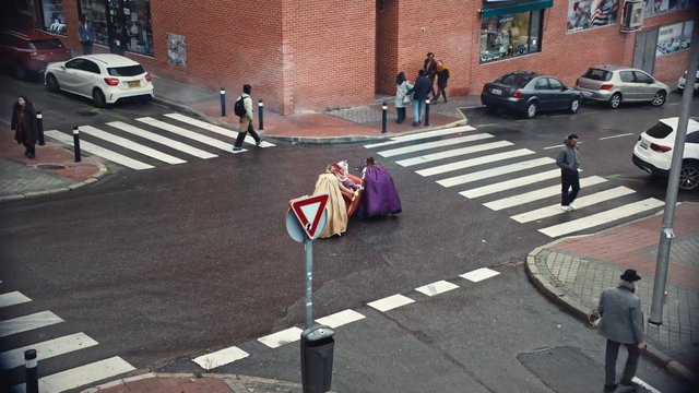 Video Reference: street, intersection, crosswalk, pedestrians, parked cars, urban, brick building, sidewalk, road sign, wet road, people group, colorful clothing, capes, city scene, outdoor, car