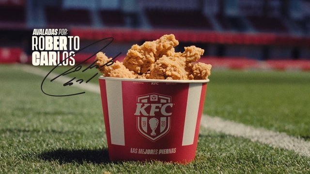 Video Reference: fried chicken, chicken bucket, fast food, sports field, grass, stadium, advertising, promotional, close-up, food, brand logo, poster, Spanish text, soccer
