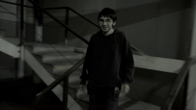 Video Reference: grayscale, indoors, staircase, railings, young man, glasses, hoodie, standing, interior, low light, black and white, industrial