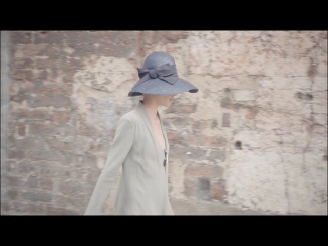 Video Reference: person, walking, hat, wide-brim hat, coat, fashion, side profile, brick wall, urban, outdoor, minimalist, pastel, motion blur