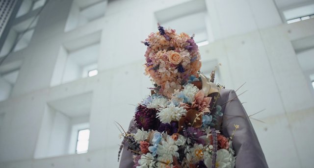 Video Reference: flower costume, floral sculpture, person, indoors, pastel colors, purple, art installation, abstract, portrait, creative, bouquet, modern architecture, low angle, decorative, fashion art