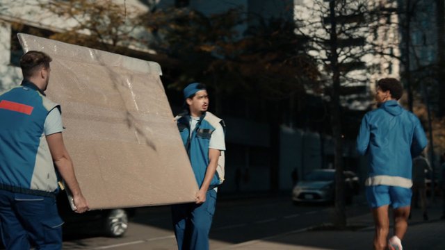 Video Reference: men, sportswear, carrying, mattress, panel, outdoor, street, moving, teamwork, blue clothing, urban, daylight, transport, work