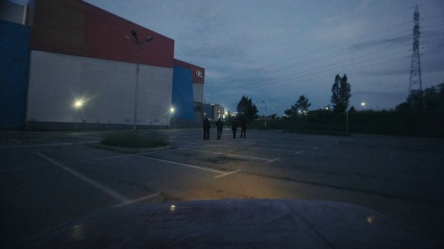 Video Reference: parking lot, dusk, industrial building, people walking, streetlights, power lines, empty lot, outdoor, evening, urban, wide shot, trees, sky, road markings