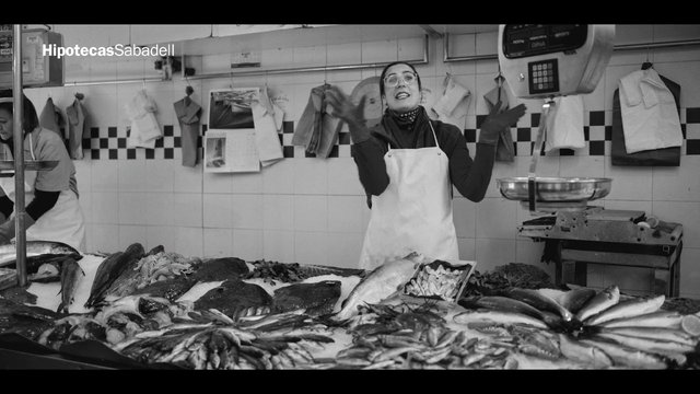 Video Reference: fish market, seafood, vendor, market stall, indoor, black and white, apron, fresh fish, scale, counter, commercial food, retail, shop interior, people, spain