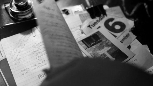 Video Reference: black and white, papers, notes, handwriting, calendar, desk, clutter, close up, blurred foreground, documents, stationery, tabletop, work area, messy, still life