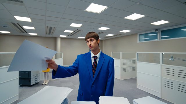 Video Reference: office, cubicles, suit, paper, business, indoor, workspace, person, blue jacket, tie, office lighting, modern office, standing, documents, cubicle partitions