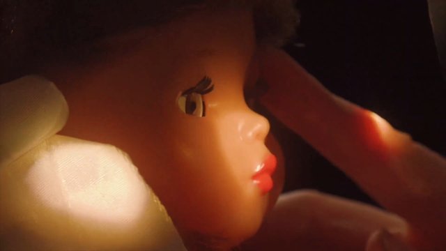 Video Reference: doll, toy, plastic, face, close-up, hand, side view, indoor, warm light, portrait, plaything, figure
