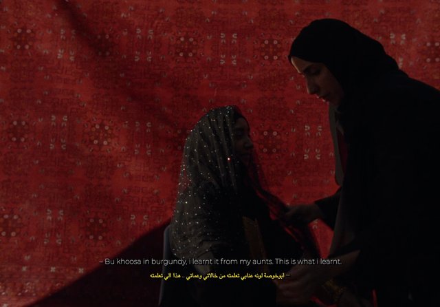 Video Reference: women, hijab, headscarf, indoor, conversation, red background, patterned fabric, dim light, close-up, subtitles, traditional clothing, standing, drama, textile