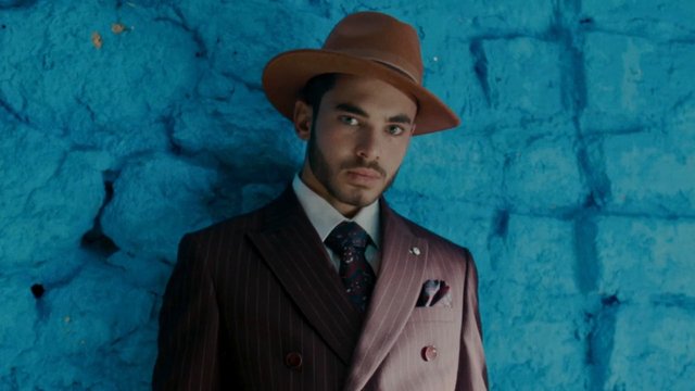 Video Reference: man, hat, suit, portrait, blue wall, fashion, formal wear, close-up, studio lighting, adult, serious expression, vintage style