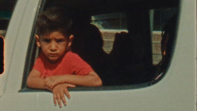 Video Reference: child, car window, vehicle, portrait, red shirt, window, travel, outdoor, leaning, serious expression, close-up, daytime