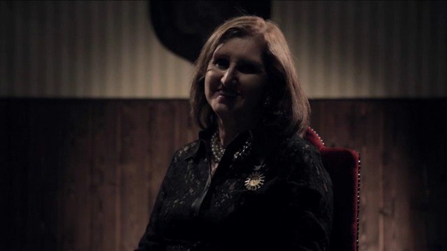 Video Reference: woman, portrait, indoor, dim light, seated, interview, wood paneling, dark background, chair, low key lighting