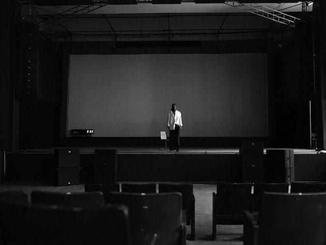 Video Reference: stage, auditorium, theater, empty seats, person, chair, speakers, dark interior, performance space, black and white, wide shot, indoor