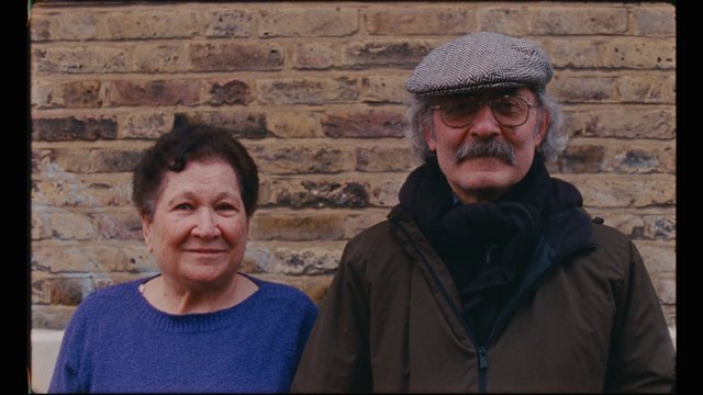 Video Reference: two people, older adults, portrait, brick wall, outdoor, posed, couple, close-up, casual clothing, hat, glasses, mustache, winter coat, medium shot, standing, background