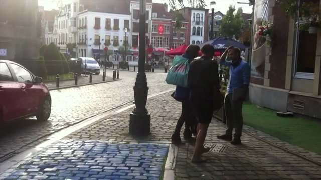 Video Reference: street, sidewalk, cobblestone, people, urban, buildings, car, outdoor, daylight, city, pedestrians, lamp post, road, intersection
