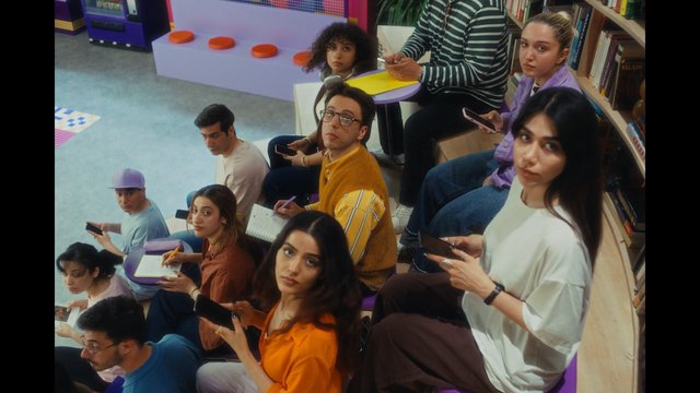 Video Reference: indoor, group, young adults, library, classroom, seated, phones, notebooks, steps, bookshelves, colorful, students, casual clothing, looking at camera