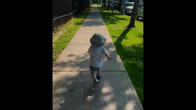 Video Reference: child, walking, sidewalk, sun hat, outdoors, daylight, suburban, trees, grass, parked cars, street, back view