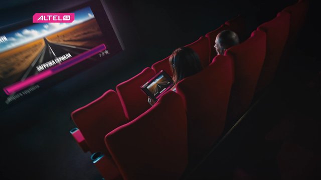 Video Reference: movie theater, red seats, audience, tablet, screen, advertisement, ALTEL 5G, dark room, cinema, people watching, technology, display, road scene, pink logo