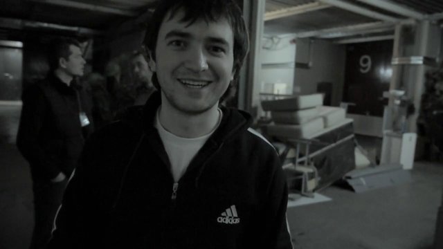 Video Reference: young man, smiling, indoor, portrait, casual clothing, group, dim light, cluttered room, close-up, black and white, standing, background person