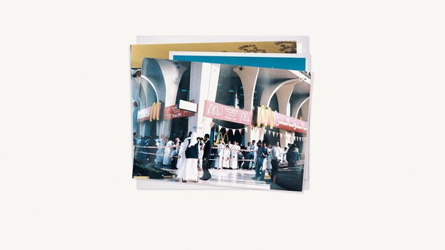 Video Reference: printed photos, photo stack, crowd, storefront, McDonald's, Arabic signage, outdoor, people, archway, retail, event, historical photo, paper, documentary, business