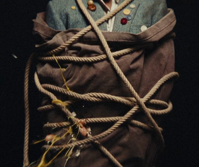 Video Reference: rope, bound, torso, jacket, coat, close-up, dark background, restraint, fabric, buttons, tied, cinematic, dramatic lighting